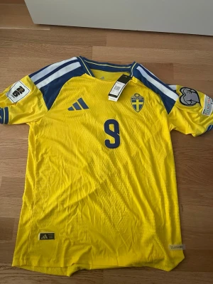 Sverige fotbollströja Isak #9 Adidas - Gul och blå officiell Sverige fotbollströja från Adidas med nummer 9 och namnet Isak på ryggen. Tröjan har blå detaljer, klubbmärke på bröstet och Climalite-material som andas. Perfekt för match eller träning. Från adidas 