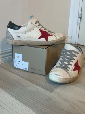 Golden Goose sneakers med röd stjärna - Snygga sneakers från Golden Goose i vitt läder med grå detaljer och en ikonisk röd stjärna på sidan. Skorna har grå snörning, svart hälparti och slitna detaljer för en cool, vintage look. Perfekta för dig som gillar streetstyle och unika skor. Priset är ej hugget i sten och går att förhandla 
