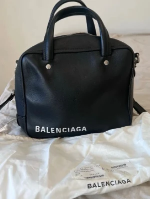 Svart Balenciaga handväska i skinn - Snygg svart handväska från Balenciaga i äkta skinn med vit logga framtill. Väskan har dubbla handtag, avtagbar axelrem och silverfärgade detaljer. Kommer med dustbag och äkthetskort. Perfekt för dig som gillar lyxiga accessoarer.