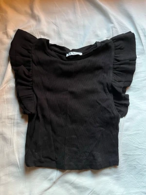 Svart ribbad topp med volangärmar - Säljer en svart ribbad topp från Zara med volangdetaljer på ärmarna. Toppen är i stretchigt material och har en rund halsringning. Perfekt för dig som vill ha något stilrent men ändå med en twist.