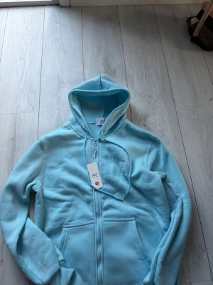 Ljusblå hoodie från Ami Paris - Snygg ljusblå hoodie från Ami Paris med dragkedja framtill och broderad logga med hjärta och A på bröstet. Tillverkad i mjuk bomullsblandning, har två fickor och justerbar huva med snören. Perfekt för en chill och stilren look. Helt ny men ganska tunnt material