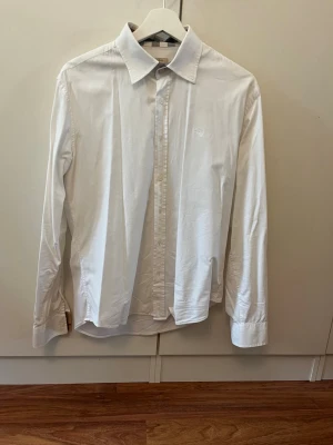 Vit skjorta från Burberry Brit - Klassisk vit skjorta från Burberry Brit med lång ärm och knappar framtill. Skjortan har en snygg krage med diskret rutigt mönster på insidan och är tillverkad i bomull. Perfekt för en stilren look.