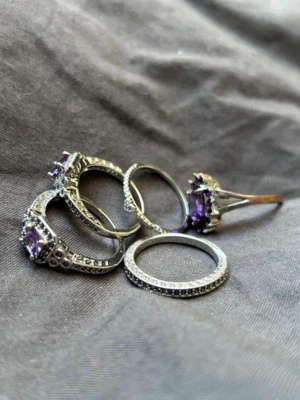 Silverringar med lila stenar - 5 stycken silver ringar, alla dekorerade med gnistrande lila stenar och små klara stenar runt om. Ringarna har olika former. Matrial stål. Storlek 18,5cm fattas en sten på ena ringen, syns på bild