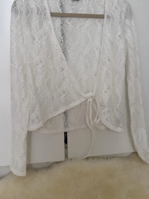 Vit spetskofta från Gina Tricot - Säljer en vit långärmad spetskofta från Gina Tricot. Koftan har knytning framtill och är gjord i ett tunt, transparent spetsmaterial med bladmönster. Perfekt att slänga över ett linne för en snygg lager-på-lager-look.