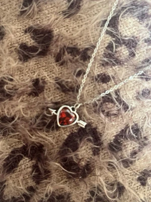 Halsband med hjärthänge och pil röd silver - Snyggt halsband i silverfärg med ett hjärthänge i röd sten och en pil som går igenom hjärtat. Kedjan är tunn och smidig, perfekt för att ge lite extra bling till din outfit. Hjärtat har en glittrande effekt som fångar ljuset snyggt. Rostfri 