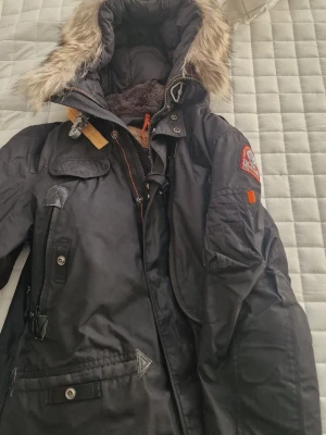 Svart parkas från Parajumpers - Svart parkasjacka från Parajumpers med avtagbar huva och fuskpäls, flera fickor och detaljer i orange. Jackan har patch på ärmen och robust dragkedja. Perfekt för kalla dagar och har en klassisk, sportig look. Säljer den billigare vid en snabb affär.