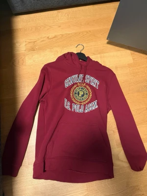 Vinröd hoodie från U.S. Polo Assn. - Använd 1 gång. Snygg vinröd hoodie med tryck 'Genuine Spirit U.S. Polo Assn.' och broderad detalj framtill. Klassisk känguruficka och dragsko i huvan. Perfekt för en avslappnad och sportig stil.