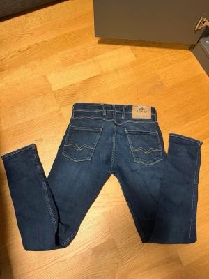 Mörkblå slim anbass jeans från Replay - Säljer ett par mörkblå skinny jeans från Replay med klassisk femficksmodell och snygga detaljer på bakfickorna. Jeansen har normal midja, smala ben och är tillverkade i stretchigt denimtyg för extra komfort. Perfekta för en trendig och avslappnad look.