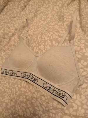 Vit Calvin Klein bralette - Vit bralette från Calvin Klein med smala axelband och elastiskt band med logga under bysten. Mjuk och bekväm modell, perfekt för en avslappnad stil. Finns en liten fläck på ena kupan vilken man kan se på bild nr 5.
