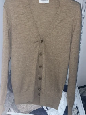 Beige kofta från Tiger Of Sweden - Stilren beige kofta från Tiger Of Sweden med v-ringning och knäppning framtill med sex bruna knappar. Långärmad och tillverkad i ett tunt, mjukt material som passar perfekt för lager-på-lager. Enkel och klassisk design som funkar till det mesta. Det finns ett litet hål som visas på bilden men den syns inte när man har på sig den.