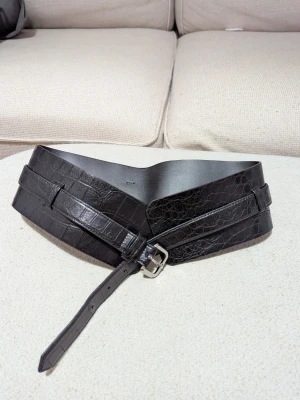 Svart brett bälte från Monki - Snyggt svart bälte från Monki i brett modell med krokopräglat mönster. Bältet har en markerad V-form framtill och silverfärgat spänne. Perfekt för att lyfta en outfit och ge midjan extra fokus.