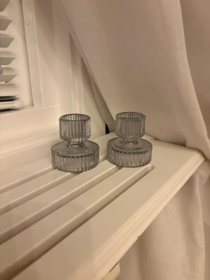 Två gråa glasljusstakar - Säljer ett par stilrena ljusstakar i grått glas med räfflad yta. De har en modern, rund form och kan användas både som ljushållare och små skålar beroende på hur de vänds. Perfekt som inredningsdetalj i hemmet.