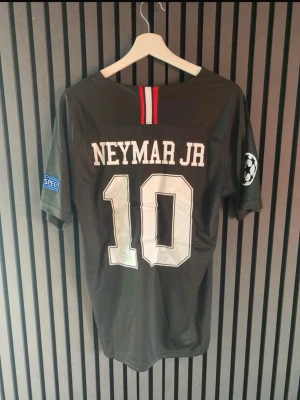 PSG Neymar Jr matchtröja svart - Säljer en svart PSG matchtröja med Neymar Jr och nummer 10 på ryggen. Tröjan har Champions League-märke på ärmen, Fly Emirates-tryck på bröstet och Paris Saint-Germain-logga. Tillverkad i lätt och ventilerande material, perfekt för fotbollsträning eller match.