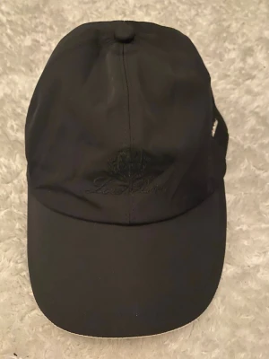 Svart keps från Loro piana  - Stilren svart keps från Loro Piana med broderad logga framtill. Klassisk böjd skärm och justerbar rem baktill. Tillverkad i ett lätt och smidigt material som passar perfekt till streetwear eller sportiga outfits.