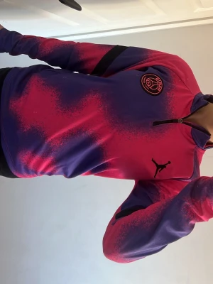 PSG träningströja rosa/lila Jordan - Säljer en snygg PSG träningströja från Jordan i rosa och lila med svart logga och emblem på bröstet. Tröjan har halv dragkedja och svart detalj på ärmen. Perfekt för fotbollsträning eller gymmet.