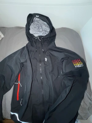 Svart vindjacka Peak Performance L - Svart vindjacka från Peak Performance med huva och dragkedja. Jackan har röda detaljer vid fickan och broderad logga på ärmen i flera färger. Innerfodret är ljust och materialet känns slitstarkt och vattenavvisande. Perfekt för blåsiga dagar.