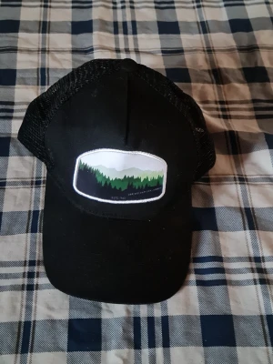 Svart truckerkeps med naturmotiv - Snygg svart truckerkeps med mesh på sidorna och ett broderat märke framtill som visar ett motiv av gröna granar och berg. Perfekt för dig som gillar outdoor-stil och vill ha en keps med coolt tryck.