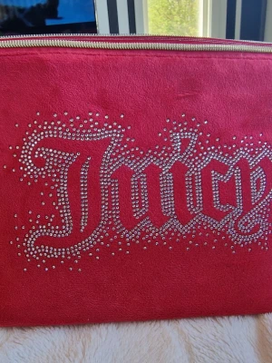Juicy Couture rosa kuvertväska - Snygg rosa kuvertväska från Juicy Couture med glittrande strasstenar som bildar texten 'Juicy' på framsidan. Väskan har en dragkedja upptill och är gjord i ett mjukt material med rosa foder inuti. Perfekt för att addera lite bling till din outfit.