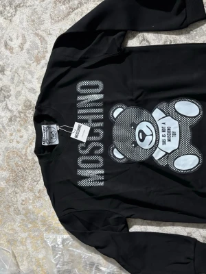 Svart Moschino sweatshirt med björn - Cool svart sweatshirt från Moschino med stort tryck av en björn och texten 'THIS IS NOT A MOSCHINO TOY' på framsidan. Tröjan har rund hals och långa ärmar. Perfekt statement-plagg för dig som gillar streetstyle och designer.