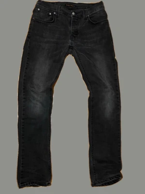 Svarta jeans från Nudie Jeans - Snygga svarta jeans från Nudie Jeans med klassisk femficksdesign och diskreta slitningar. Jeansen har raka ben och subtila detaljer på bakfickorna. Perfekta för dig som gillar en stilren look.