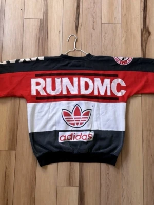 Adidas RUN DMC färgglad hoodie - Snygg och unik hoodie från Adidas med RUN DMC-tryck i rött, svart och vitt. Framsidan har en stor stadssilhuett och logga, baksidan har RUN DMC-tryck och Adidas-märke. Perfekt för dig som gillar streetwear och retrostil.