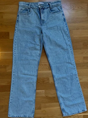 Ljusblå jeans med paljetter från ZARA - Snygga ljusblå raka jeans från ZARA med massor av glittrande strass över hela framsidan. Klassisk femficksmodell med hög midja och bälteshällor. Perfekta för dig som vill sticka ut och addera lite extra bling till din outfit.