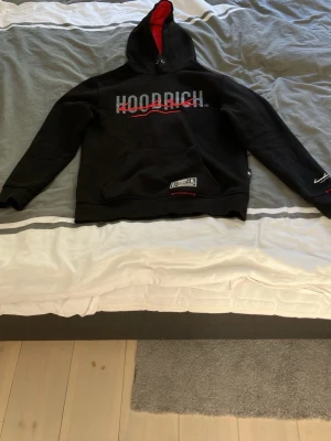 Svart HOODRICH hoodie med röd detalj - Svart hoodie från Hoodrich med stor vit och röd logga på bröstet och tryck på ryggen. Insidan av huvan är röd, vilket ger en snygg kontrast. Klassisk känguruficka framtill och ribbade muddar. Perfekt för en streetwear-look. Är storlek XS men om man har någon större storlek så passar den nog lite bättre.