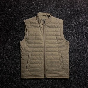 Grå quiltad dunväst från Riley - Stilren grå dunväst från Riley i storlek L. Västen har quiltad design, hög krage och dragkedja framtill. Perfekt att slänga över en hoodie eller tröja för extra värme. Finns liten fläck men syns inte mycket, kolla sista bilden. Två fickor på sidorna och diskret look. Hör av er vid minsta fundering!