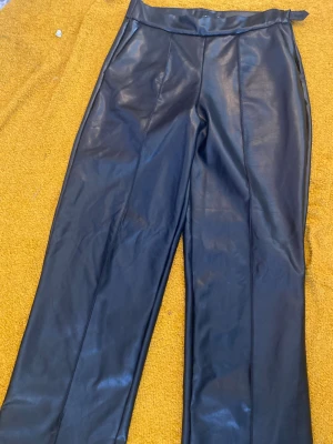 Svarta skinn byxor  - Stilrena svarta skinnbyxor  med hög midja och raka ben. Byxorna har en snygg glansig finish och markerad söm framtill. Perfekta för dig som gillar en trendig och clean look.