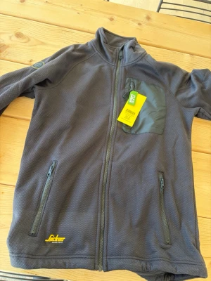 Svart fleecejacka från Snickers Workwear - Helt ny aldrig använd. Snygg svart fleecejacka från Snickers Workwear med hel dragkedja, två sidofickor med dragkedja och en bröstficka. Jackan har hög krage och diskret logga på bröstet. Perfekt för lager-på-lager och passar till både jobb och fritid. 
