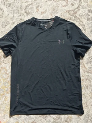 Svart tränings-tshirt från Under Armour - Svart t-shirt från Under Armour med diskret logga på bröstet och tryck längs sidan. Tillverkad i ett lätt och snabbtorkande polyestermaterial, perfekt för träning. Klassisk passform med korta ärmar och rund hals.