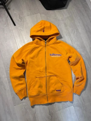 Gul Peak Performance hoodie med dragkedja - Gul hoodie från Peak Performance med lila broderad logga och text på bröstet. Modellen har dragkedja framtill, huva och känguruficka. Tillverkad i mjukt bomullsmaterial och har ribbade muddar vid ärmslut och nederkant.