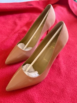 Nude Kate Heels 100mm från Christian Louboutin - Säljer ett par klassiska nude pumps från Christian Louboutin i glansigt lackmaterial. Skorna har spetsig tå och hög klack på 100mm, vilket ger en elegant och tidlös look. Perfekta för dig som vill ha ett par stilrena och lyxiga klackar. Dessa skor är även helt oanvända!