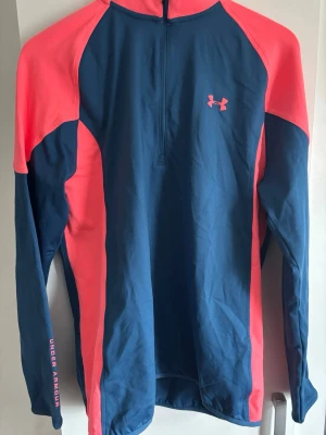 Under Armour golftröja blå/korall - Sportig långärmad träningströja från Under Armour i blått och korallrosa. Tröjan har halv dragkedja framtill, logga på bröstet och ärm, samt tryck på ryggen. Perfekt för löpning eller gym. Materialet är lätt och andas.