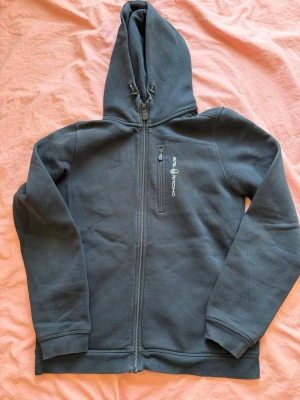 Mörkblå hoodie från Sail Racing - Mörkblå hoodie med dragkedja från Sail Racing. Har en bröstficka med dragkedja och logga tryckt i vitt på bröstet. Justerbar huva med snören och ribbade muddar vid ärmslut och nederkant. Perfekt för en chill och sportig look.