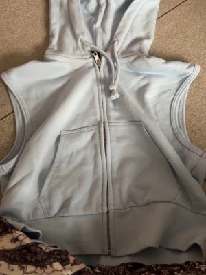 Ljusblå ärmlös hoodie från H&M Divided - Säljer en ljusblå ärmlös hoodie från H&M Divided. Hoodien har dragkedja framtill, stor känguruficka och justerbar huva med snören. Perfekt för lager-på-lager och chill stil. Tillverkad i mjukt bomullsmaterial.