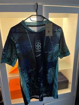 Nike Brasil träningströja blå mönstrad - Snygg blå träningströja från Nike med Brasil-logga på bröstet. Tröjan har tropiskt blad- och djurmönster i olika blå och turkosa nyanser, rund hals och korta ärmar. Perfekt för fotbollsträning eller gymmet. Materialet är lätt och andas.