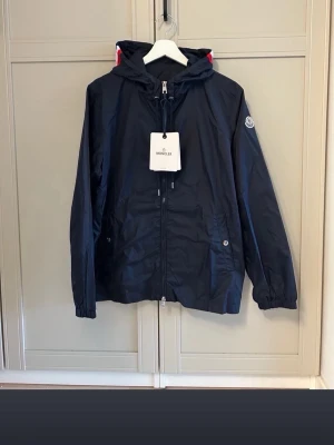 Marinblå vindjacka från Moncler - Stilren marinblå vindjacka från Moncler med huva och dragkedja framtill. Jackan har vita och röda detaljer på huvan, två fickor med knapp och Moncler-logga på ärmen. Perfekt för blåsiga dagar och enkel att matcha. Ingår påse och tags osv.