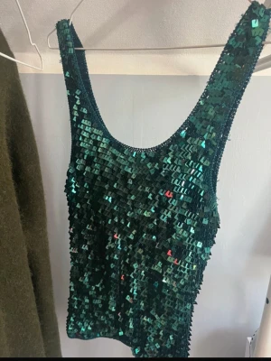 Grön paljettlinnetopp - Snygg grön linne-topp täckt med glittriga paljetter. Modellen är ärmlös med breda axelband och rundad halsringning. Perfekt för dig som vill sticka ut och glänsa lite extra. Aldrig använd!