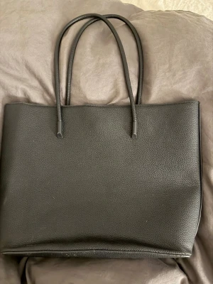 Svart stilren handväska i konstläder - Snygg svart handväska i konstläder med enkel och stilren design. Väskan har två långa handtag och stängs med magnetknapp. Insidan är rymlig med en innerficka för småsaker. Perfekt för dig som gillar minimalistisk look.