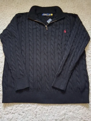 Svart kabelstickad tröja Polo Ralph Lauren - Kabelstickad svart tröja från Polo Ralph Lauren med halv dragkedja och ribbad krage. Tröjan har det klassiska röda Polo-logot broderat på bröstet och ribbade muddar vid ärmslut och nederkant. Perfekt för lager på lager och snygg till jeans.