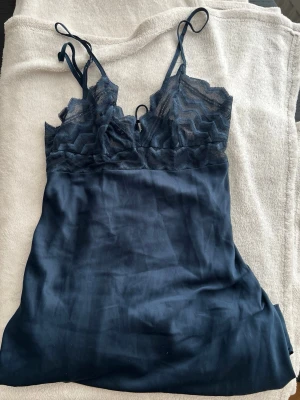 Mörkblå spetsnattlinne med smala band - Ny pris 699kr. Snyggt mörkblått nattlinne med tunna axelband och spetsdetaljer upptill. Överdelen har ett vågigt spetsmönster och dekorativ snörning framtill. Materialet känns mjukt och följsamt, perfekt för en avslappnad kväll hemma.