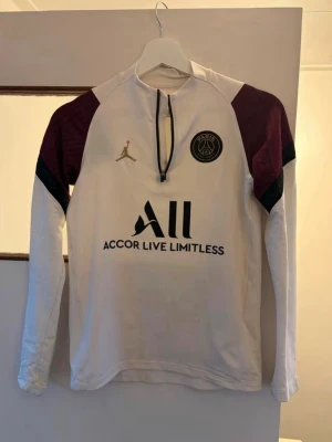 PSG långärmad träningströja vit/bordeaux - Snygg långärmad träningströja från PSG och Jordan i vitt med bordeauxfärgade axelpartier och svarta detaljer. Tröjan har halv dragkedja, klubbmärke på bröstet och tryckt sponsorlogga. Perfekt för fotbollsträning eller streetwear.