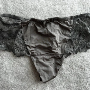 Trosa Missya spetstring - Säljer ett par grå stringtrosor med breda spetsdetaljer på sidorna och en liten rosett framtill. Trosorna har en mjuk och stretchig känsla med elegant spetsmönster som ger en feminin look. Perfekta för dig som gillar stilrena underkläder med fina detaljer.