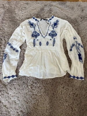 Vit blus med blå brodyr, Zara - Supersnygg vit blus med blå broderade detaljer och v-ringning. Blusen har långa ärmar och ett lätt, luftigt tyg som ger en bohemisk känsla. Perfekt att styla med jeans eller kjol för en trendig look till sommaren. Aldrig använt 🌸