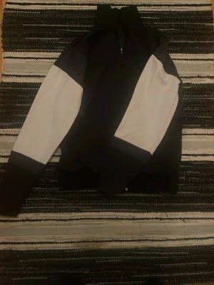 Svart och vit vindjacka med dragkedja - Snygg svart och vit vindjacka med hög krage och hel dragkedja framtill. Jackan har kontrasterande vita partier på ärmarna och en sportig look. Perfekt för dig som gillar streetwear och vill ha en stilren jacka till vardags.