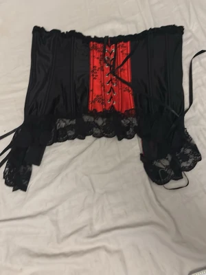 Svart och röd korsett med spets - Snygg korsett i svart och rött med snörning framtill och dekorativa spetsdetaljer längs nederkanten. Korsetten har blommigt mönster på de röda partierna och breda band för att justera passformen. Materialet är glansigt tyg och spets.