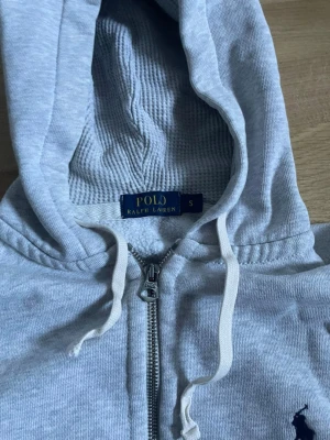 Grå hoodie från Polo Ralph Lauren - En grå zip-up hoodie från Polo Ralph Lauren med en stilren och tidlös design. Tillverkad i ett mjukt och bekvämt material som passar perfekt för vardagligt bruk. Den har en smidig dragkedja framtill och är lätt att matcha med olika outfits. Passar bäst för personer som är cirka 170 - 175 cm långa och ger en snygg, bekväm passform.