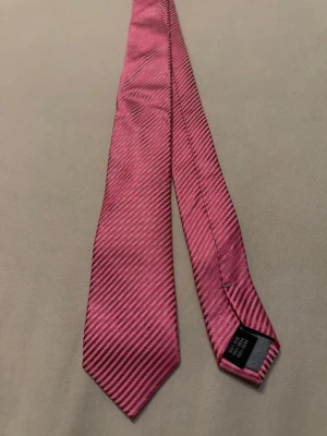 Rosa slips med smala ränder - Säljer en snygg rosa slips med tunna diagonala ränder i en lite mörkare nyans. Slipsen har en klassisk form och är tillverkad i ett glansigt material som ger en stilren look. Perfekt för dig som vill addera lite färg till outfiten.