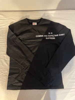 Svart långärmad Supreme x Comme des Garçons - Svart långärmad topp från Supreme och Comme des Garçons med vit text på bröstet. Klassisk crewneck och normal passform. Tillverkad i mjuk bomull, perfekt för en clean streetwear-look.